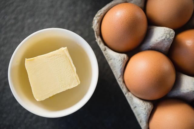This image shows a close-up view of a bowl with a piece of butter next to a carton of brown eggs on a table. It can be used for culinary blogs, recipe websites, cooking tutorials, and nutritional articles. The image highlights essential baking ingredients, making it suitable for content related to baking, cooking, and kitchen preparations.