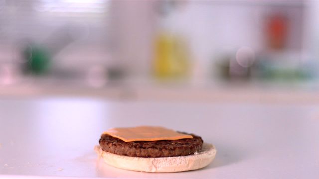 Shows a slice of cheese falling onto a beef patty placed on a bun in a kitchen setting. Ideal for illustrating culinary techniques, food preparation, or fast food cooking. Perfect for blogs or articles on burgers, kitchen gadgets that aid in cooking, or advertisements related to food products.