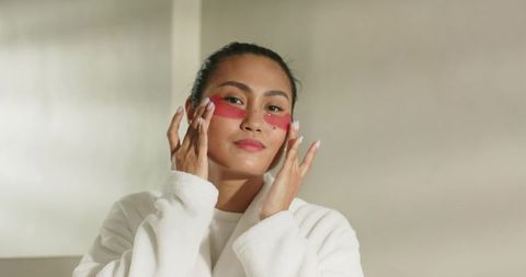 Mid-adult asian woman applying red under-eye patches wearing white robe for skincare glow