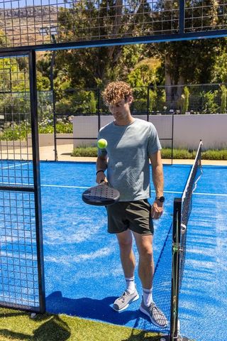 Young man standing on vivid blue padel court holding paddle and bouncing ball on sunny day