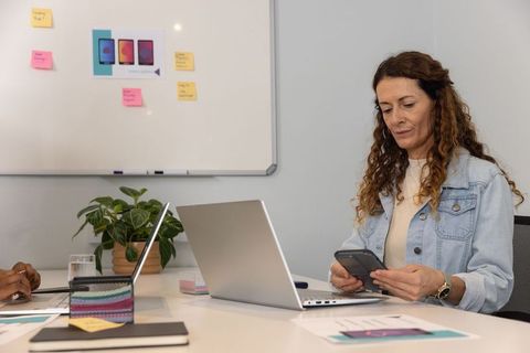Mature professional checking smartphone while using laptop in collaborative meeting room