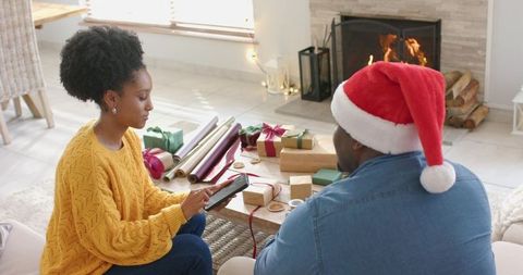 Couple wrapping christmas gifts by fireplace using tablet, cozy holiday home scene