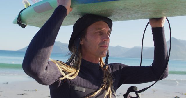 Man with dreadlocks wearing a wetsuit and hat carries surfboard on his shoulder on a sunny beach. Mountains and clear blue water in background. Ideal for travel ads, surfing events, outdoor activities, adventure blogs, and lifestyle promotions.