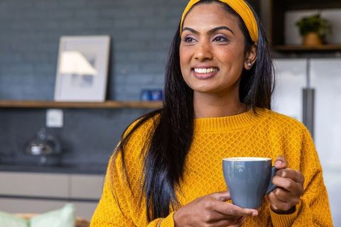 Indian woman in her thirties enjoying coffee in modern kitchen wearing mustard sweater