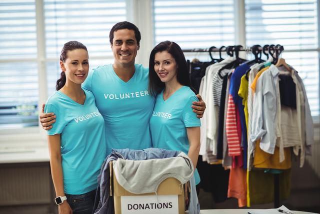 Three smiling volunteers standing together in a workshop, organizing clothing donations. They are wearing matching blue shirts with 'Volunteer' printed on them. This image is ideal for promoting community service, charity events, nonprofit organizations, and volunteer recruitment campaigns.