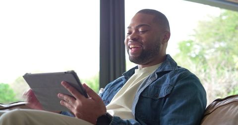 Smiling man relaxing on sofa using tablet at home in daylight wearing denim jacket