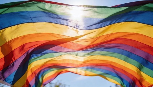 Backlit rainbow pride flag billowing in sunlight over blue sky and treetops