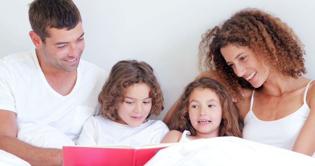 Family consisting of parents and two children reading a book together while sitting in bed. All members of the family are smiling and engaging in the activity, creating a heartwarming scene of togetherness and bonding. This image can be used in promotions related to family life, bedtime routines, or educational content for children.