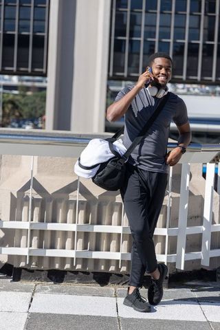 Smiling african american man leaning on rooftop railing talking on phone with gym bag