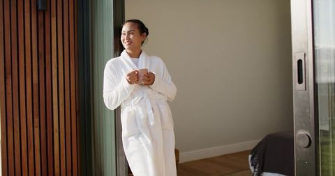 Asian woman standing in doorway wearing white bathrobe holding mug smiling in morning sun