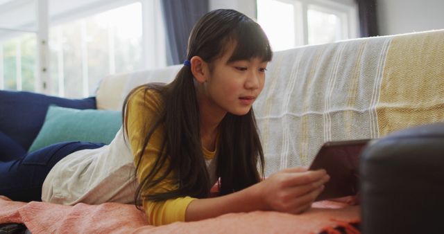 Teenage girl lies on a comfortable sofa, reading an eBook on her tablet during a casual afternoon at home. Perfect for depicting home leisure, technology use, and youthful relaxation. Ideal for use in advertisements, website banners, and social media posts related to technology, lifestyle, and home comfort.