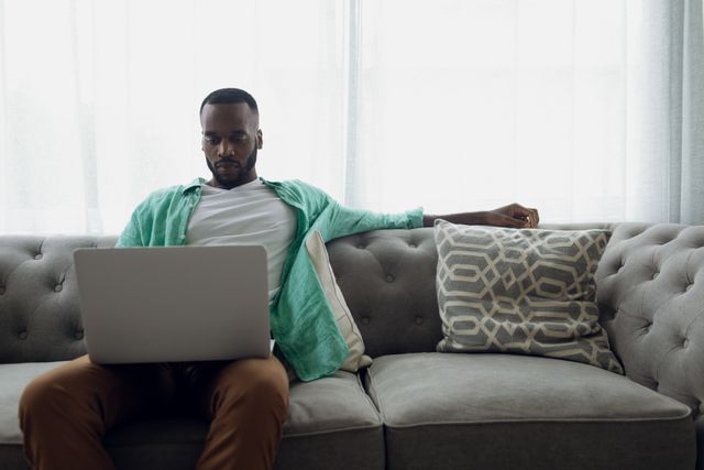 Front view of an African-American man sitting on a grey couch with white curtains inside a room while using a laptop