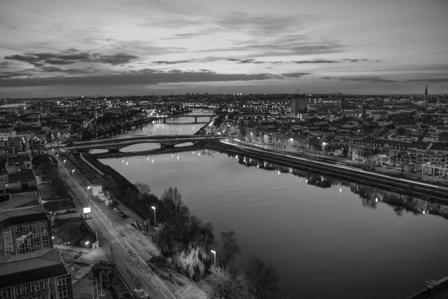 This black and white image captures a beautiful cityscape at dusk, showcasing a river flowing through the center and lit-up bridges. Urban skyline with softly twinkling city lights and a calm, reflective river can provide a sophisticated aesthetic to any design. Perfect for city-themed websites, backgrounds for presentations, or editorial content focused on urban living or photography.