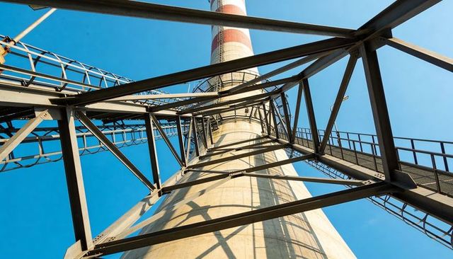 Rising industrial chimney framed by steel trusses and ladders against clear blue sky