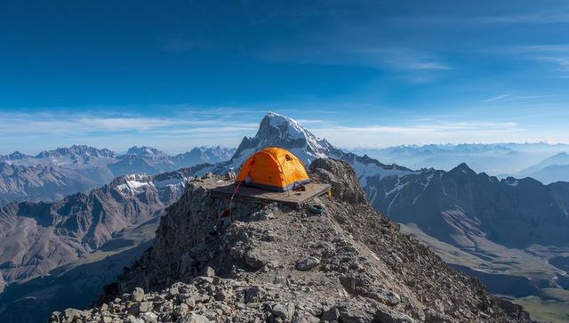 Camping on narrow summit ridge with orange dome tent overlooking snowy alpine peaks