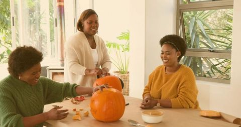 Three women carving pumpkins in bright kitchen celebrating autumn gatherings