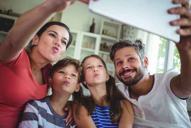 A happy family of four is taking a selfie at home using a digital tablet. The parents and two children are smiling and making playful faces, capturing a moment of togetherness and fun. This image can be used for promoting family-oriented products, technology, lifestyle blogs, or advertisements focusing on family bonding and modern living.