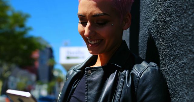 Young woman with short pink hair smiling while looking at her smartphone. She is standing against a dark wall outdoors on a sunny day, wearing a leather jacket and facial piercings. Ideal for use in ads and articles on modern lifestyle, fashion, technology use, and outdoor activities.