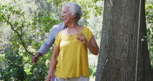 Elderly couple walking together in a sunlit forest, enjoying nature and each other's company. The elderly woman is laughing while her partner follows closely. Suitable for use in advertisements promoting active aging, senior lifestyle, health and wellness, retirement living, and nature therapy. Ideal for illustrating the joy of spending quality time outdoors in retirement.