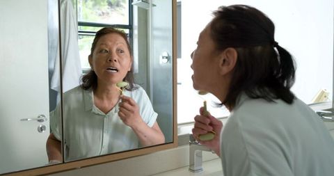 Senior asian woman performing jade roller facial massage while checking mirror in bathroom
