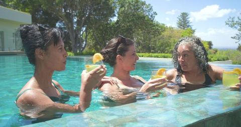 Three women relaxing in villa pool sipping cocktails on sunny vacation