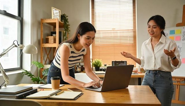 Asian coworkers collaborating at sunny creative office desk with laptop and notebooks