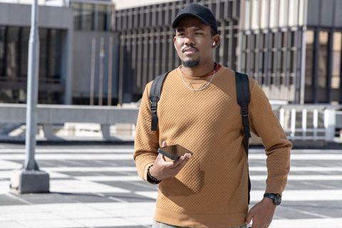 Young african american commuter standing in urban plaza holding smartphone and backpack
