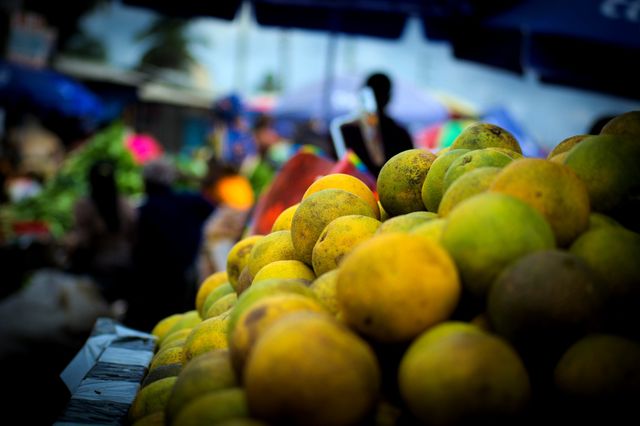 Bright bunches of fresh oranges are piled up at an outdoor local market stall, showcasing vibrant tropical fruit ready to be purchased. Lively and colorful background with other produce and market activity visible, highlighting a bustling local market atmosphere. This image can be used to depict fresh produce, healthy eating, market environments, or organic vendors.