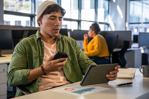 Asian man using tablet and smartphone at modern open-plan office desk viewing chart
