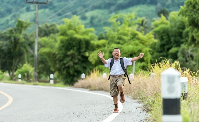 Boy running energetically on a rural road wearing a school uniform and backpack. The lush green scenery and winding road emphasize the joys of childhood and the importance of education. Suitable for themes related to childhood happiness, outdoor activities, rural lifestyle, or educational content.