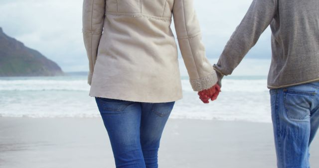 A young Caucasian couple holds hands while walking along a scenic beach, with copy space. Their casual attire and the tranquil coastal setting suggest a romantic and peaceful moment together.
