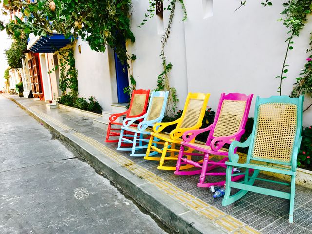 Colorful rocking chairs lined up along a white wall on an urban sidewalk. Each chair is painted a different bright color, creating a vibrant and cheerful scene. Ideal for use in content about urban life, street decor, outdoor seating, leisure activities, and vibrant environments.