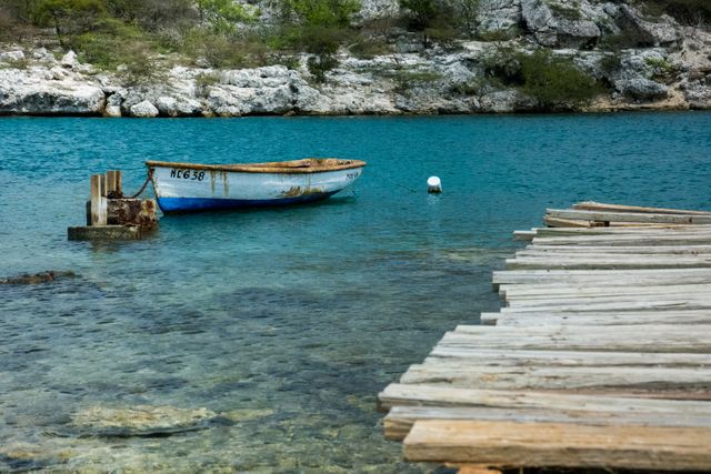 Serene scene of a single wooden rowboat tied near a weathered wooden dock on clear turquoise water with a rocky shoreline in the background. Perfect for illustrating themes of tranquility, nature, and lakeside living. Useful for travel brochures, nature magazines, outdoor lifestyle blogs, and serene relaxation prints.