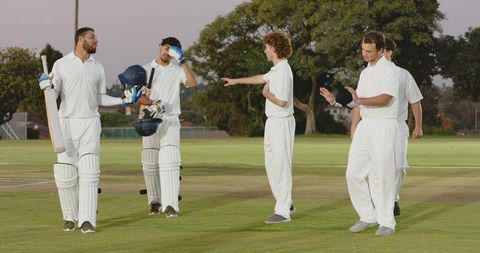 Diverse male cricketers walking off pitch at dusk wearing white kit holding bats and helmets
