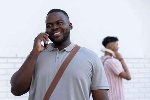 Smiling black man talking on smartphone with friend in background, casual urban commute