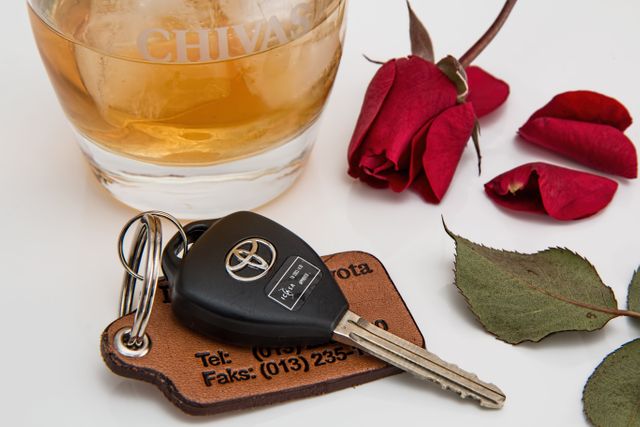Car key beside filled whiskey glass and scattered red rose petals on a white table. Image can be used for promoting romantic occasions, vehicle branding, responsible drinking messages, mixed messages about drinking and driving, or as contemplative artistic imagery.