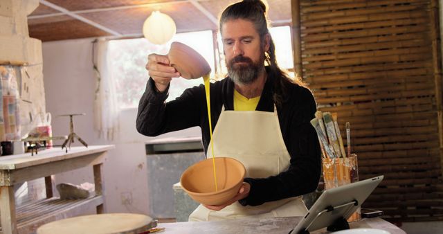 Bearded Pottery Artist Pouring Yellow Glaze from Bowl in Rustic Studio - Download Free Stock Images Pikwizard.com