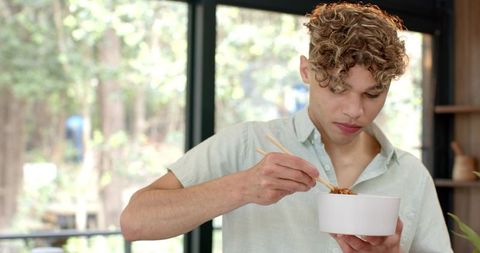 Young man enjoying noodles with chopsticks in bright kitchen