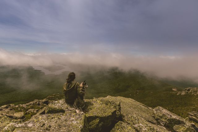 Person with dog sitting on rocky cliff edge, looking at expansive mountain valley beneath a cloudy sky. Useful for themes of adventure, solitude, nature exploration, documentation of beautiful landscapes, or companionship between humans and pets.
