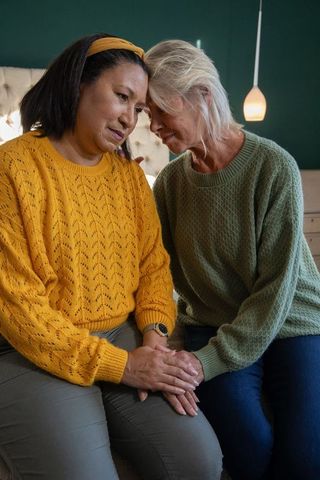 Two senior women holding hands on bed offering comfort and emotional support