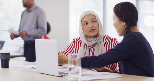 Businesswomen discussing project while sitting at desk in modern office. One wears a hijab, symbolizing diversity and inclusion. Colleagues working in background add to a professional environment. Useful for business articles, corporate websites, and diversity initiatives.