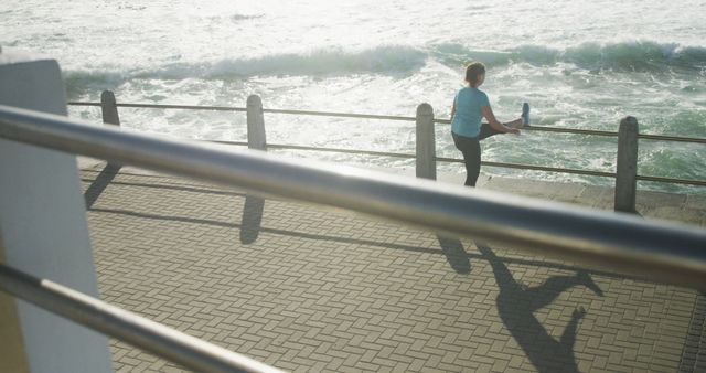 Woman leaning against railing, stretching leg, beside ocean during a sunny morning. Great for fitness and wellness articles, outdoor workout promotions, or healthy lifestyle campaigns. Emphasizes importance of exercising outdoors and maintaining an active lifestyle.