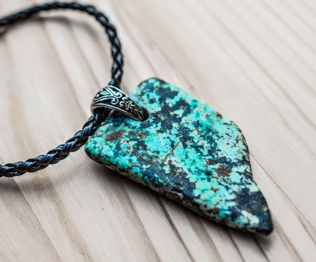 Close-up of a handmade turquoise stone arrowhead pendant necklace with ornate design on a braided leather cord, lying on a wooden surface. Ideal for designers showcasing bohemian or rustic fashion accessories, online jewelry stores highlighting unique and handcrafted pieces, or bloggers discussing distinctive fashion items and accessories.