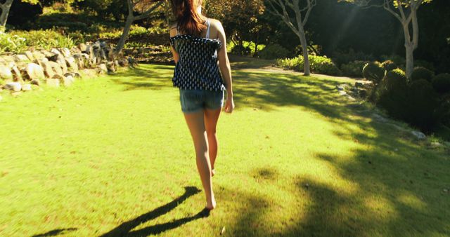Image shows a woman walking barefoot on a sunny lawn in a park. She wears a blue polka-dotted blouse and denim shorts. The park is filled with green grass, trees, and some rocks in the background, with sunlight casting shadows. This image can be used in websites or blogs focusing on leisure activities, nature walks, summer fashion, or mental relaxation.