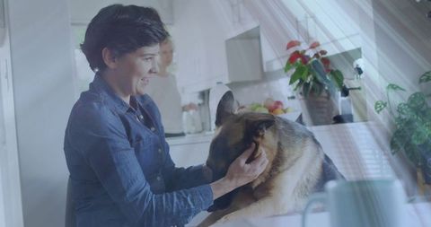 Smiling woman cradling german shepherd head in sunlit kitchen with turquoise mug, showing pet affect