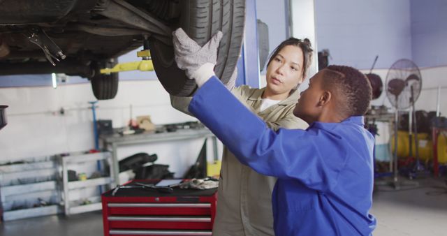 Female mechanic guiding young apprentice on changing car tire in an auto workshop. Useful for content related to automotive training, professional mentoring, workshop skills, and vocational education in the automotive industry. Image can also support themes around empowering female professionals and encouraging youth education in trades.