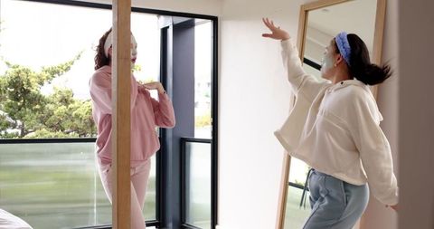 Two friends enjoying skincare routine and dancing by full-length mirror in sunlit bedroom