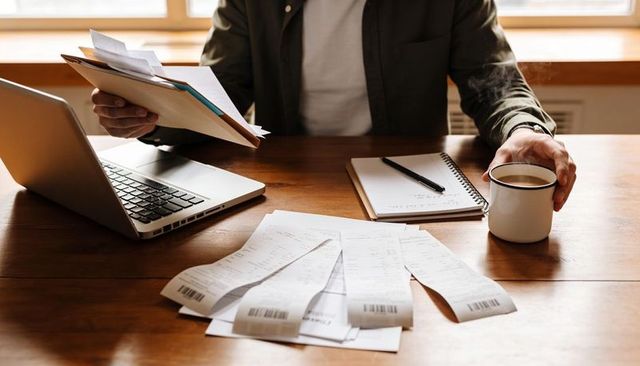 Man reviewing receipts and bills at home desk with laptop and coffee cup