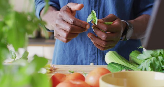 Close-up of a person preparing fresh vegetables, showcasing hands picking herbs over a wooden countertop. Tomatoes and various greens are in focus, emphasizing organic and healthy food preparation at home. Ideal for use in culinary blogs, healthy lifestyle promotions, and cooking tutorials.