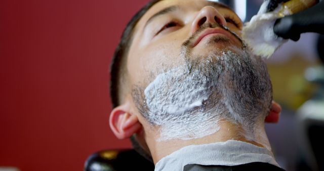 A close-up view of a man at a barber shop receiving a shaving foam application on his beard. Suitable for use in articles or advertisements focusing on men's grooming, barber services, relaxation techniques, and modern masculinity. Ideal for blogs, websites, and promotional materials for barbershops or grooming products.